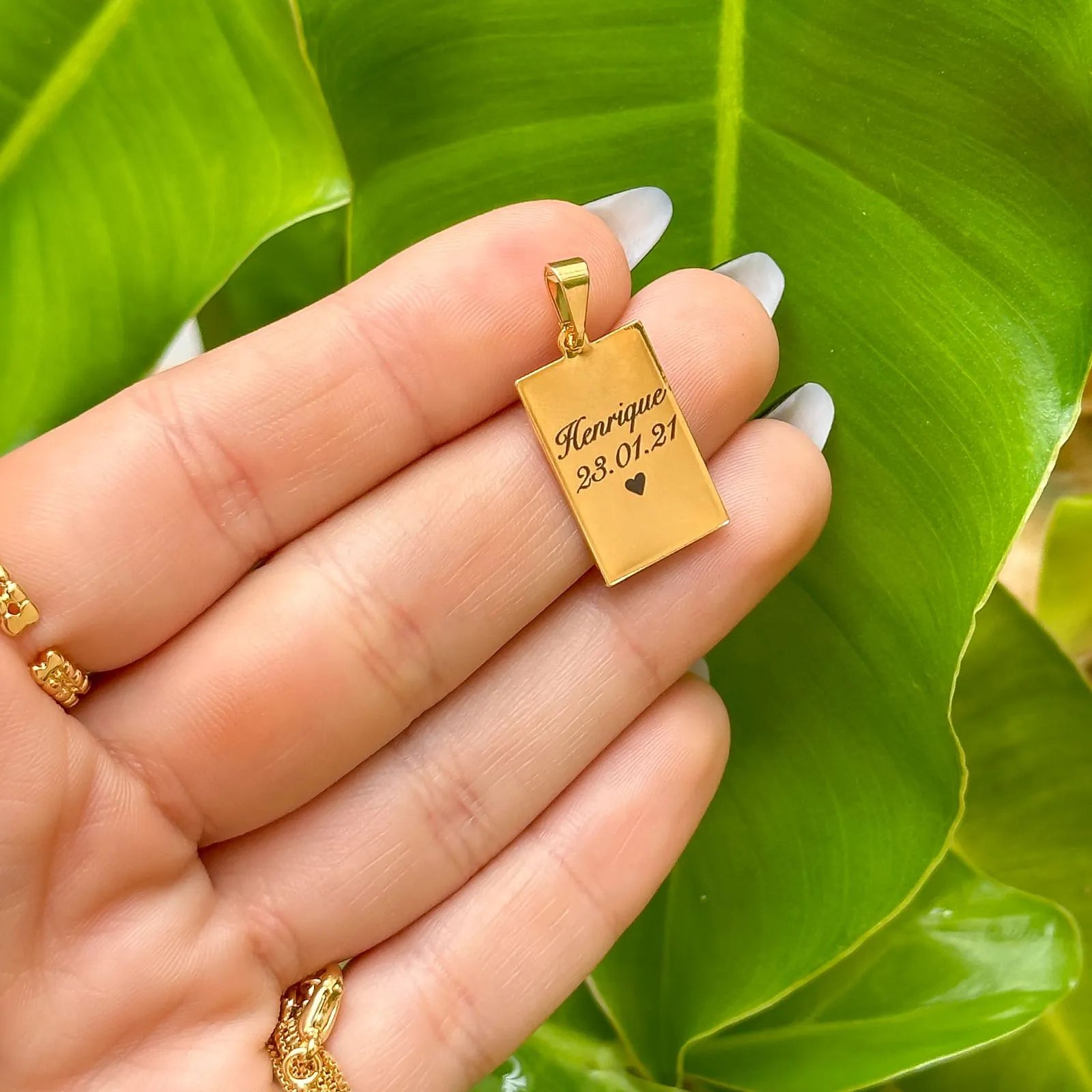 Pingente retangular dourado personalizado com nome e data sendo exibido na mão